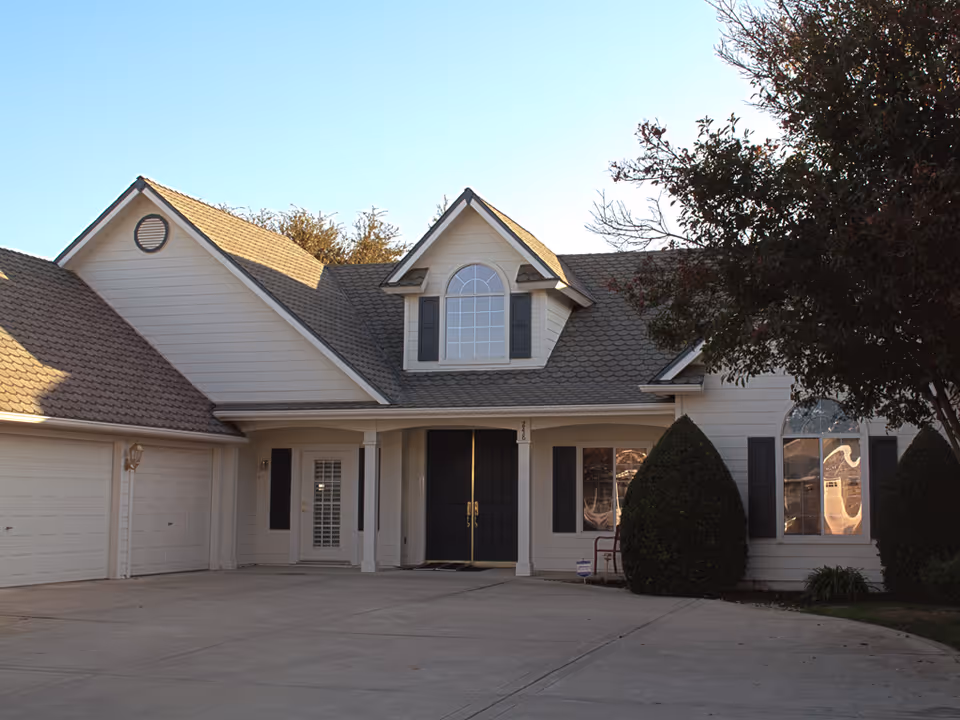 Front exterior of a residential-looking building with a double garage, central covered entrance, dormer window, and trimmed shrubs.