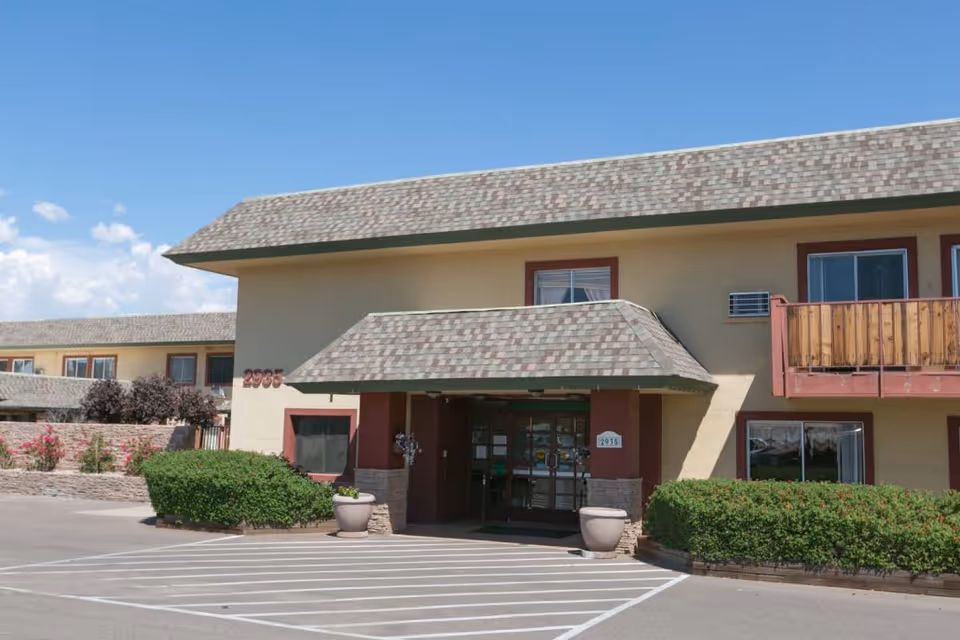 Exterior view of Rose Court Senior Living building with a beige facade, a shingled roof, and an entrance covered by a small awning. There are bushes and flower pots near the entrance, and a clear blue sky in the background.