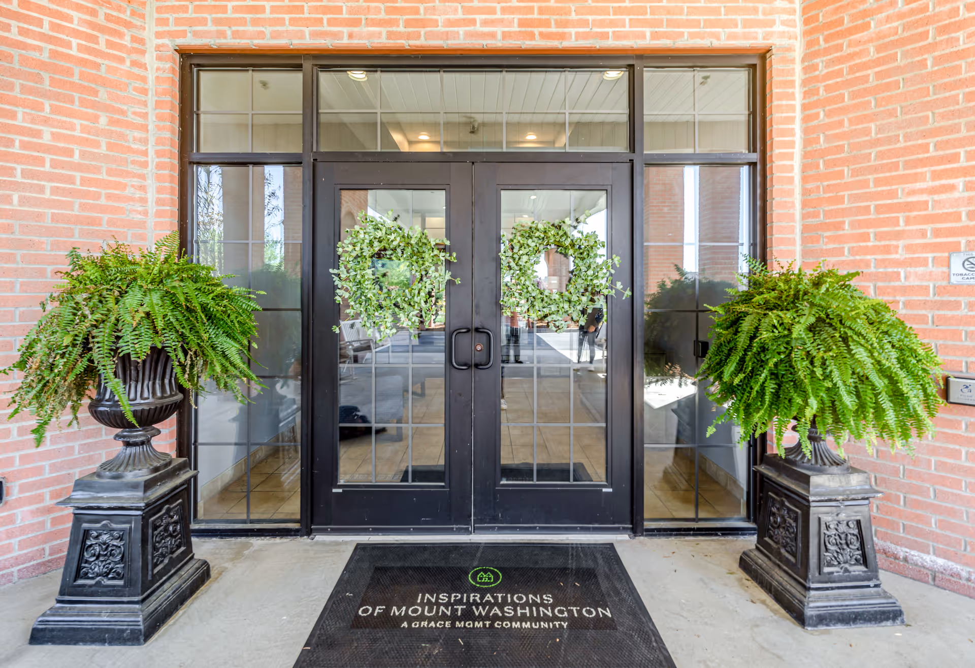 Entrance to Inspirations of Mount Washington facility with double glass doors decorated with green wreaths, flanked by two large potted ferns on ornate black pedestals, and a black welcome mat displaying the facility name.