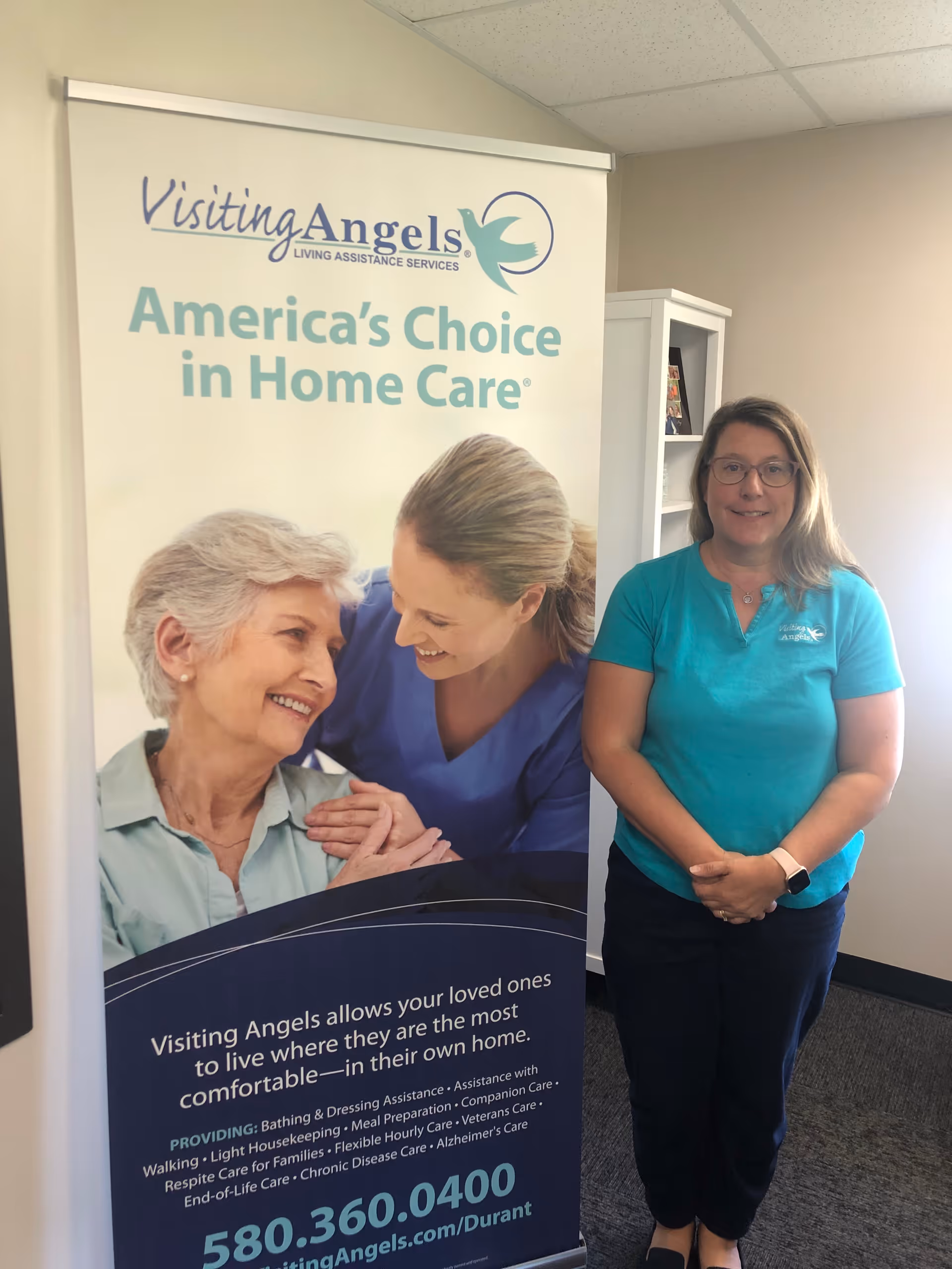 Staff member in teal polo stands beside a Visiting Angels banner indoors.