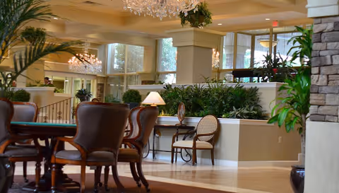 Spacious hotel-style lounge with chandeliers, plants, and cushioned chairs in a bright lobby.