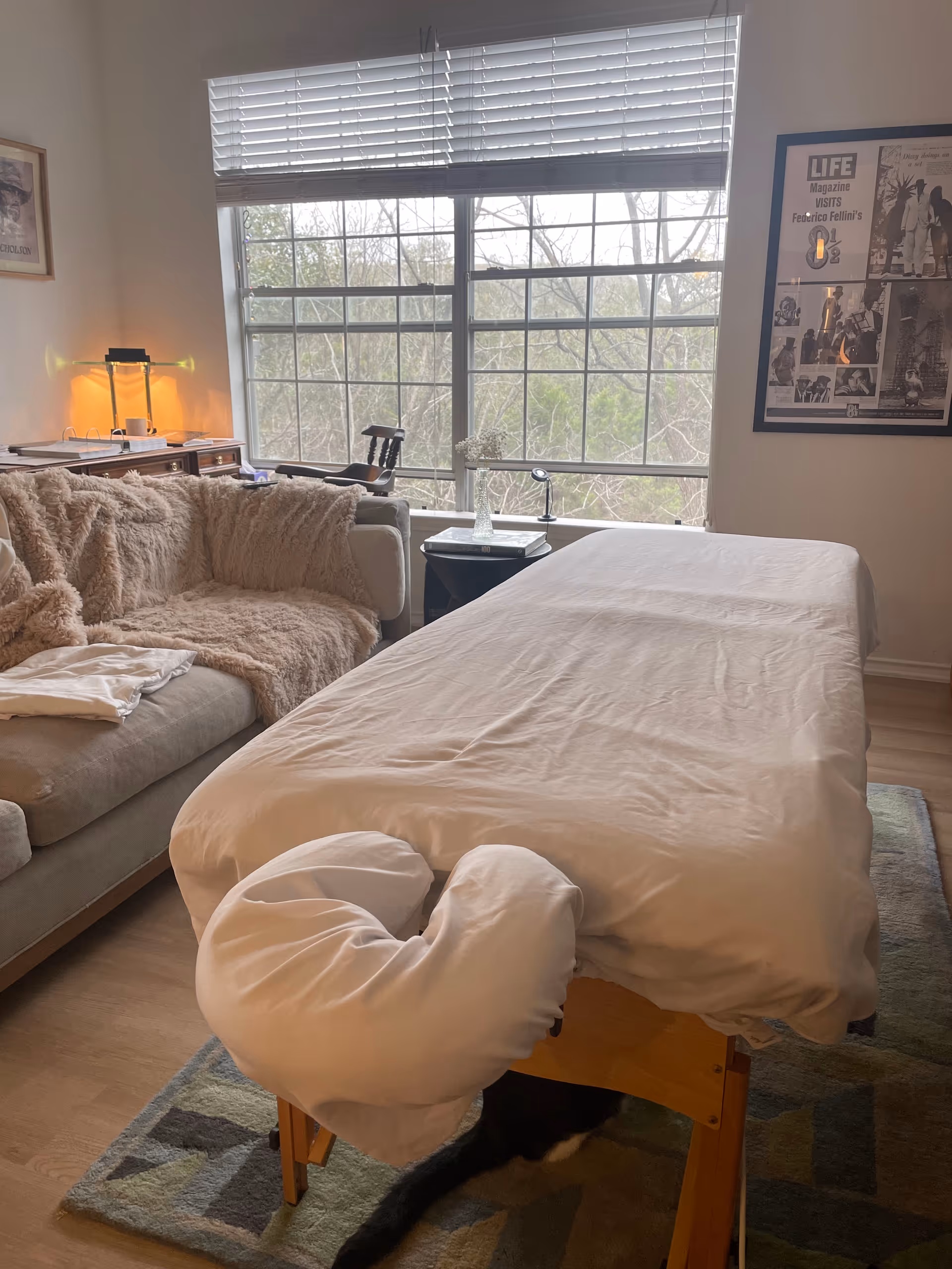 A massage table covered in white sheets sits in a cozy living room with large window.