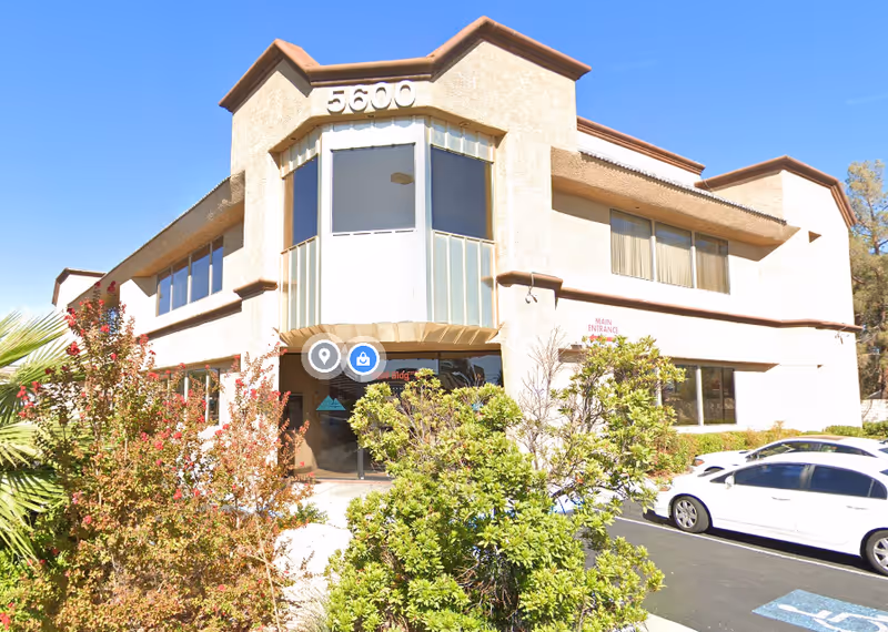 Exterior view of a beige two-story care facility with a glass entrance and parked cars