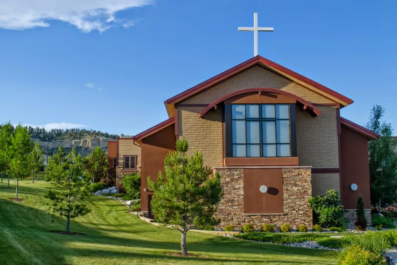 Exterior view of a building with a large cross on the roof, surrounded by green grass, small trees, and shrubs under a blue sky with some clouds.