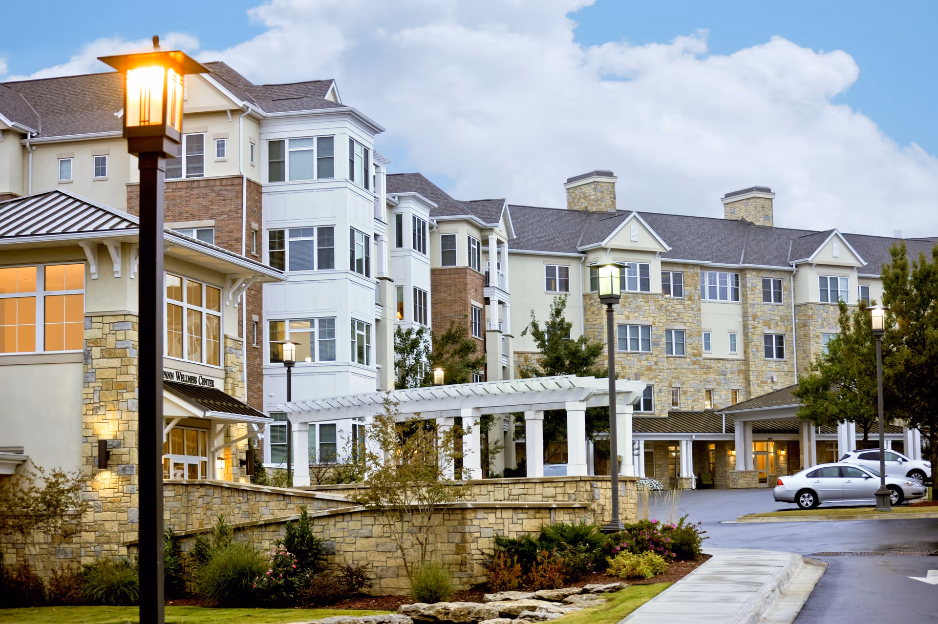 Exterior view of a multi-story senior living facility building with stone and beige siding, large windows, a covered entrance, and a well-maintained landscaped area with shrubs and flowers. Several street lamps are lit along the driveway, and a few cars are parked near the entrance under a cloudy sky.
