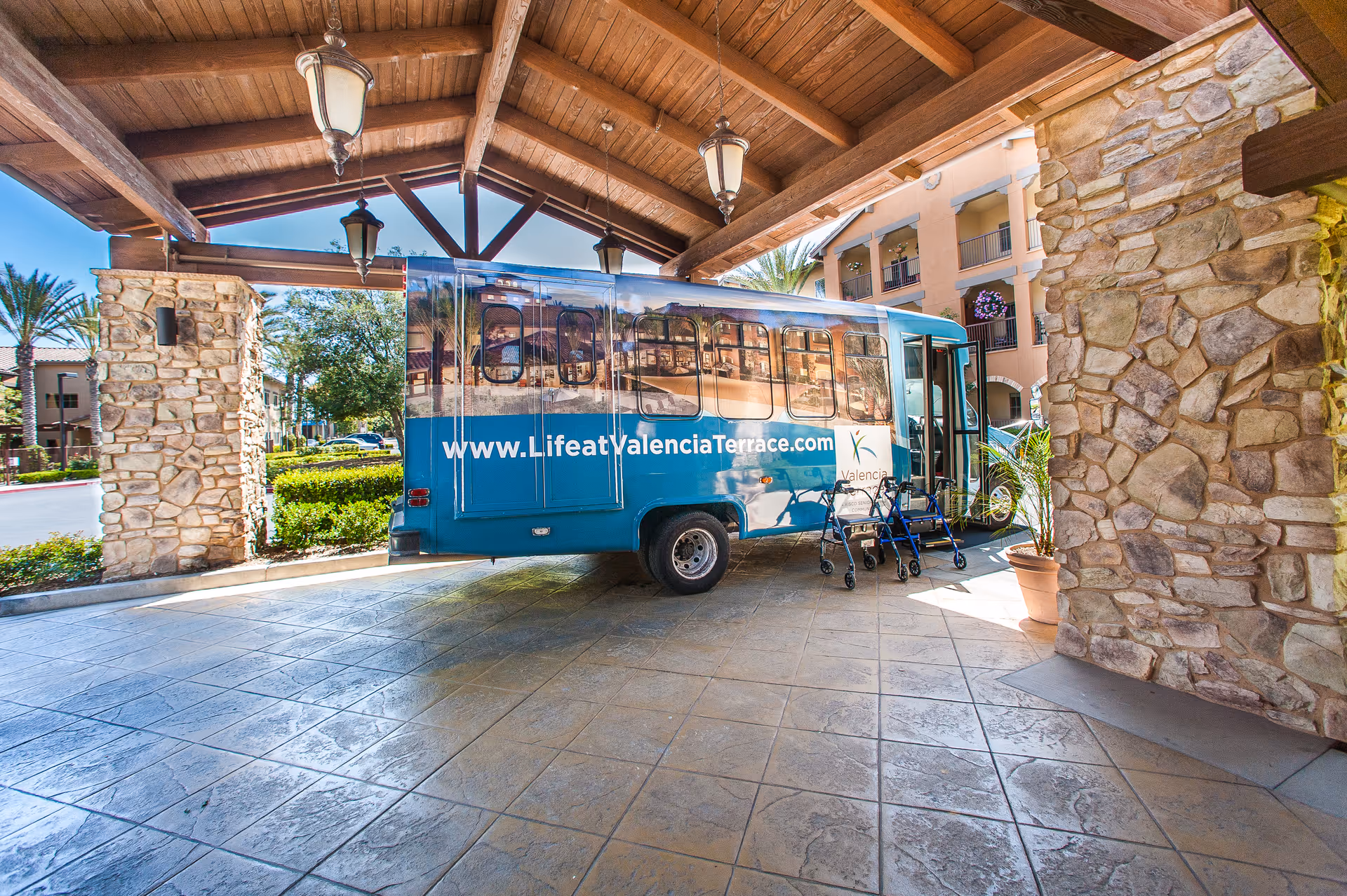 A blue shuttle bus parked under a wooden roofed entrance area with stone pillars. The bus has the website www.LifeatValenciaTerrace.com and the Valencia Terrace logo on its side. Two walkers are placed near the bus door. The background shows part of the building and some greenery.