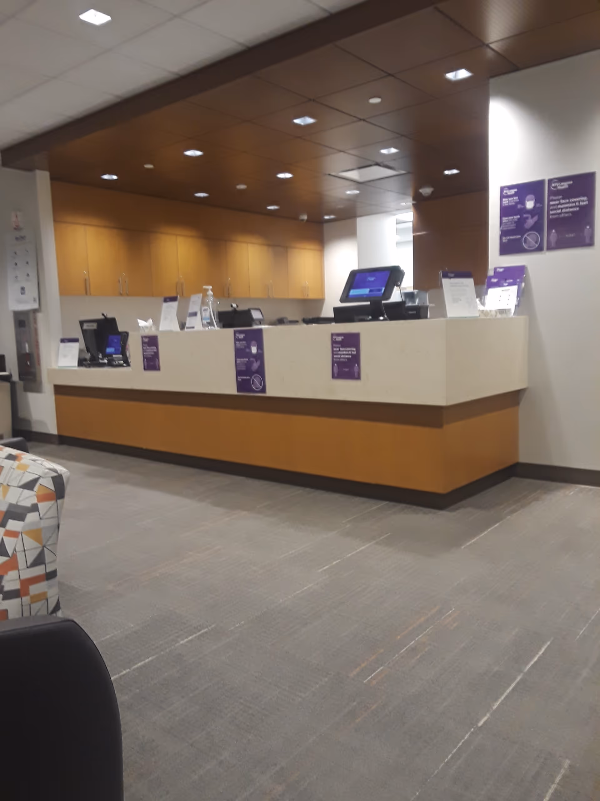 Interior hospital lobby with long beige reception desk, computers, wood cabinets, and purple posters.