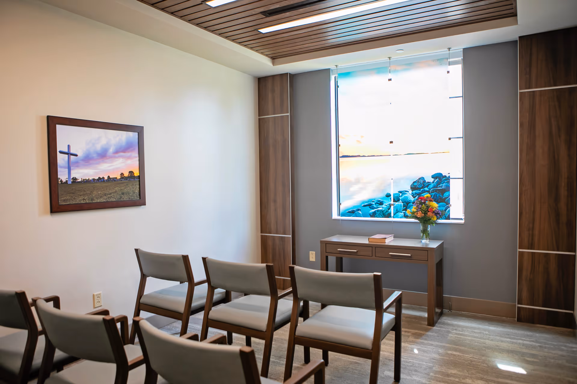 A small room with six wooden chairs arranged in two rows facing a wooden table. On the table, there is a vase with colorful flowers and a closed book. The wall behind the table features a large window with an image of a rocky shoreline and water. Another wall has a framed picture of a cross at sunset. The room has a modern ceiling design with wooden slats.