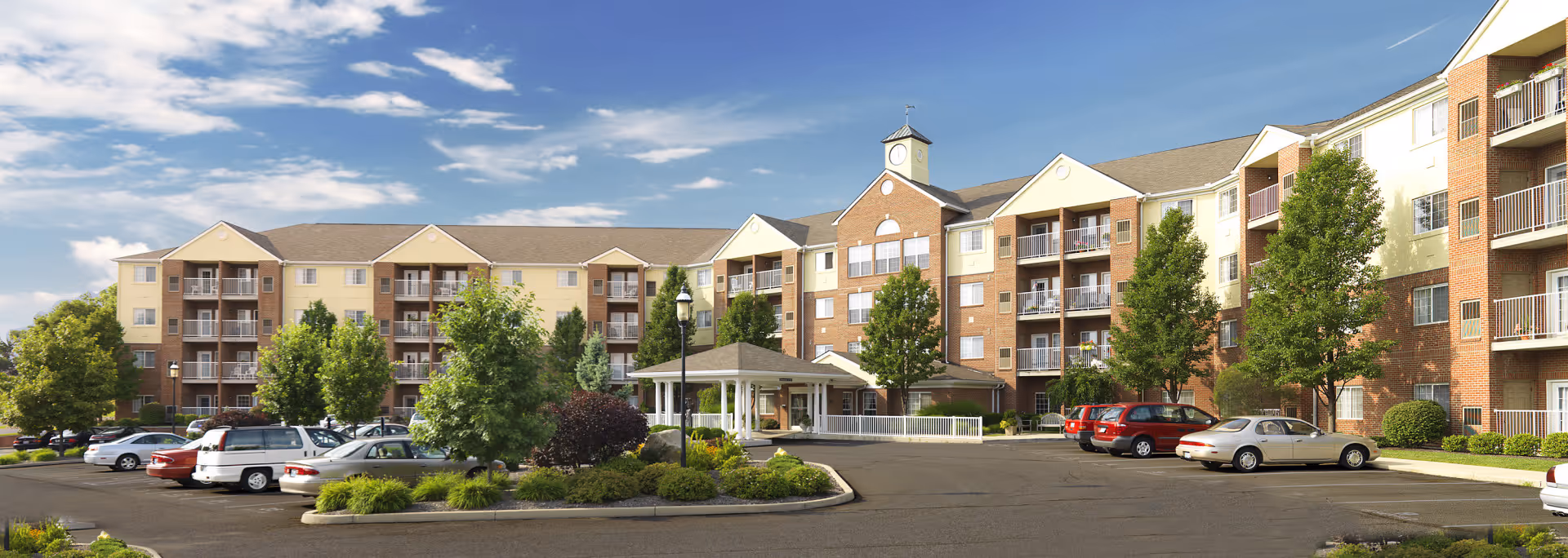 Exterior view of a large, multi-story senior living facility building with balconies, surrounded by trees and a parking lot with several cars parked. The sky is partly cloudy with blue patches.