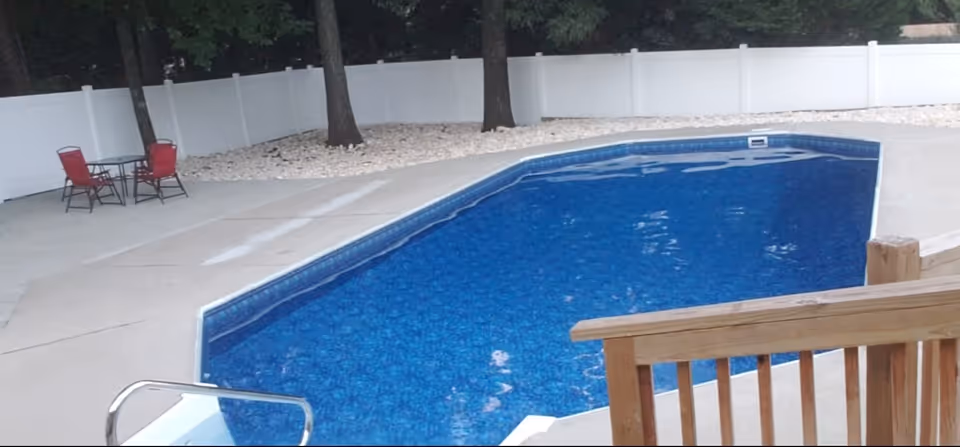 Outdoor swimming pool with blue water, concrete deck, white fence, and red patio chairs.