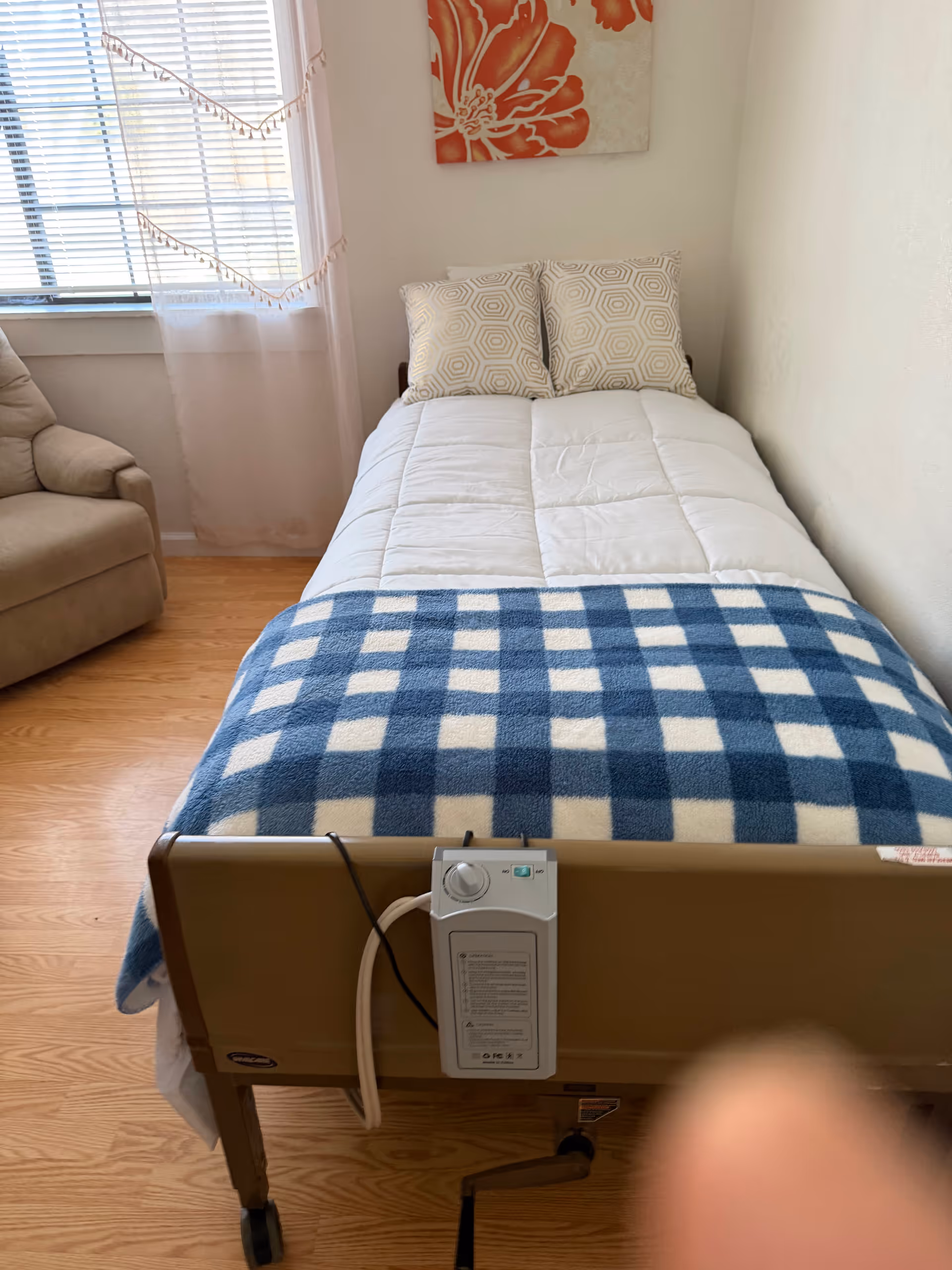 A bedroom with a single bed, blue plaid blanket, two pillows, beige chair, and window.