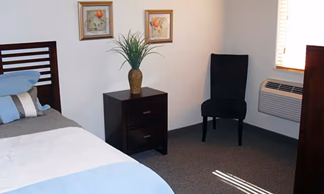 A simple bedroom with a bed, nightstand topped with a vase of greenery, a chair, wall art, and a window air conditioner.