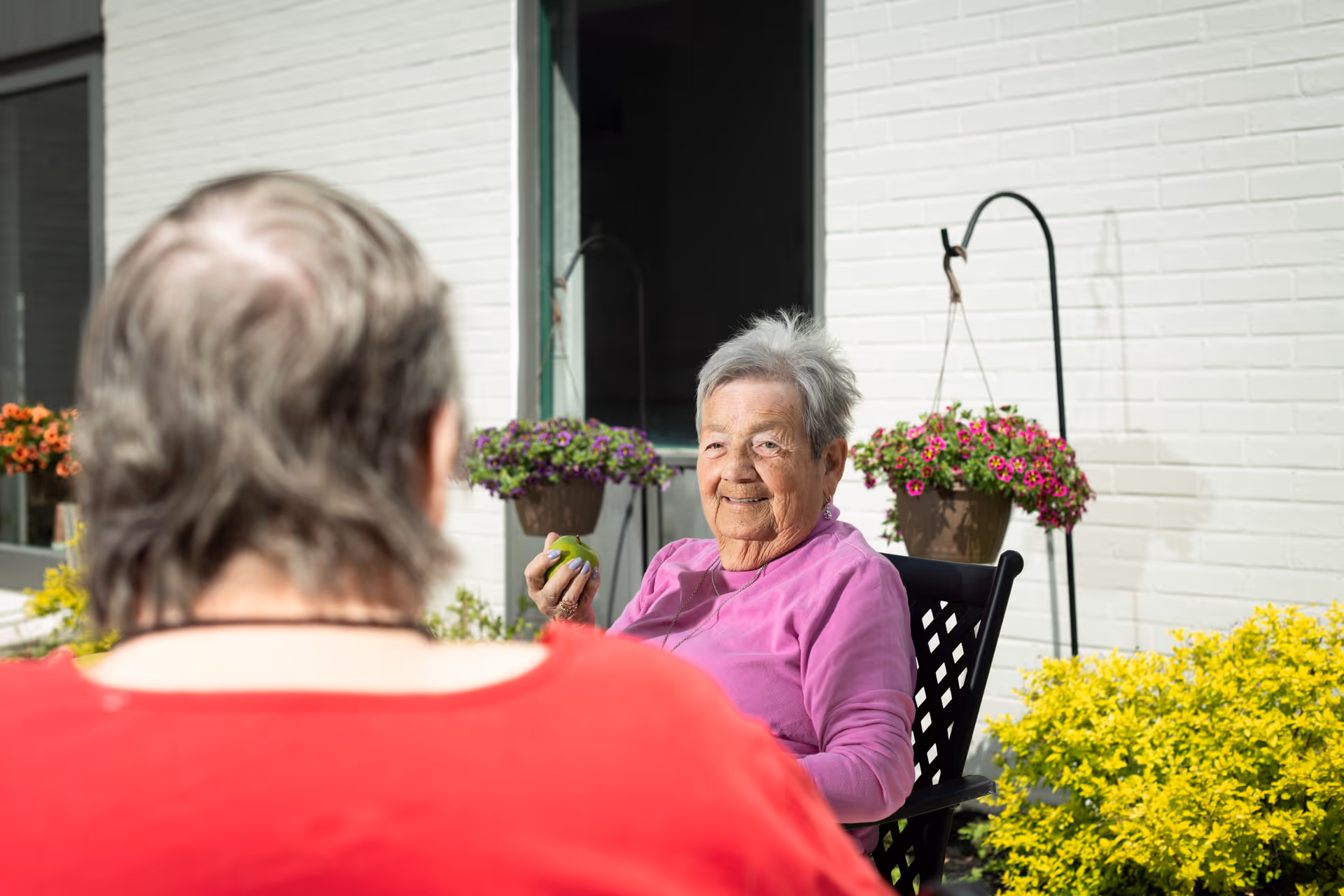 Two elderly women sitting outside near a white brick building. One woman, wearing a pink top, is smiling and holding a green apple. The other woman, seen from behind, is wearing a red top. There are hanging flower pots with purple and pink flowers and yellow bushes nearby.