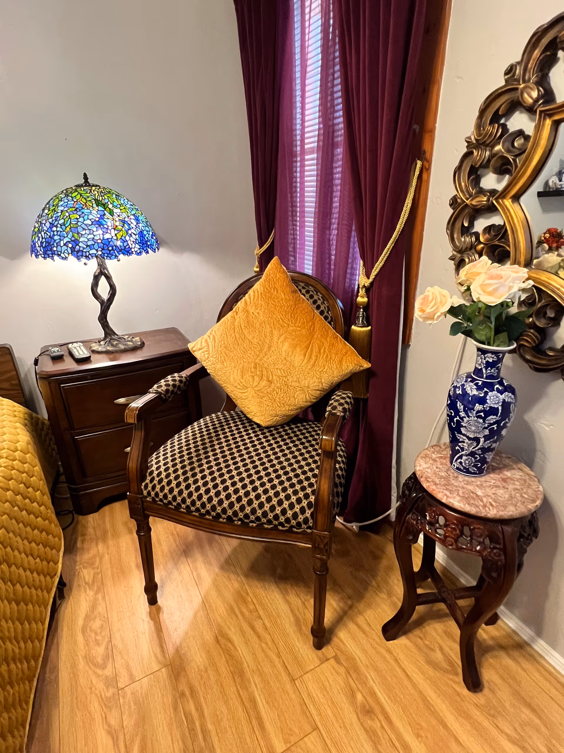 A cozy corner of a room featuring a wooden armchair with patterned upholstery and a mustard yellow cushion. Next to the chair is a wooden nightstand with a colorful stained glass lamp and remote controls on top. Behind the chair, there are burgundy curtains with gold tiebacks covering a window. To the right, a small ornate wooden table holds a blue and white vase with light pink roses, and a decorative gold-framed mirror hangs on the wall above it. The floor is light wood.