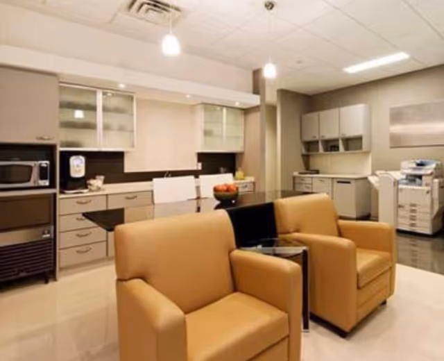 Modern communal lounge and kitchenette with tan armchairs, cabinets, and office equipment.