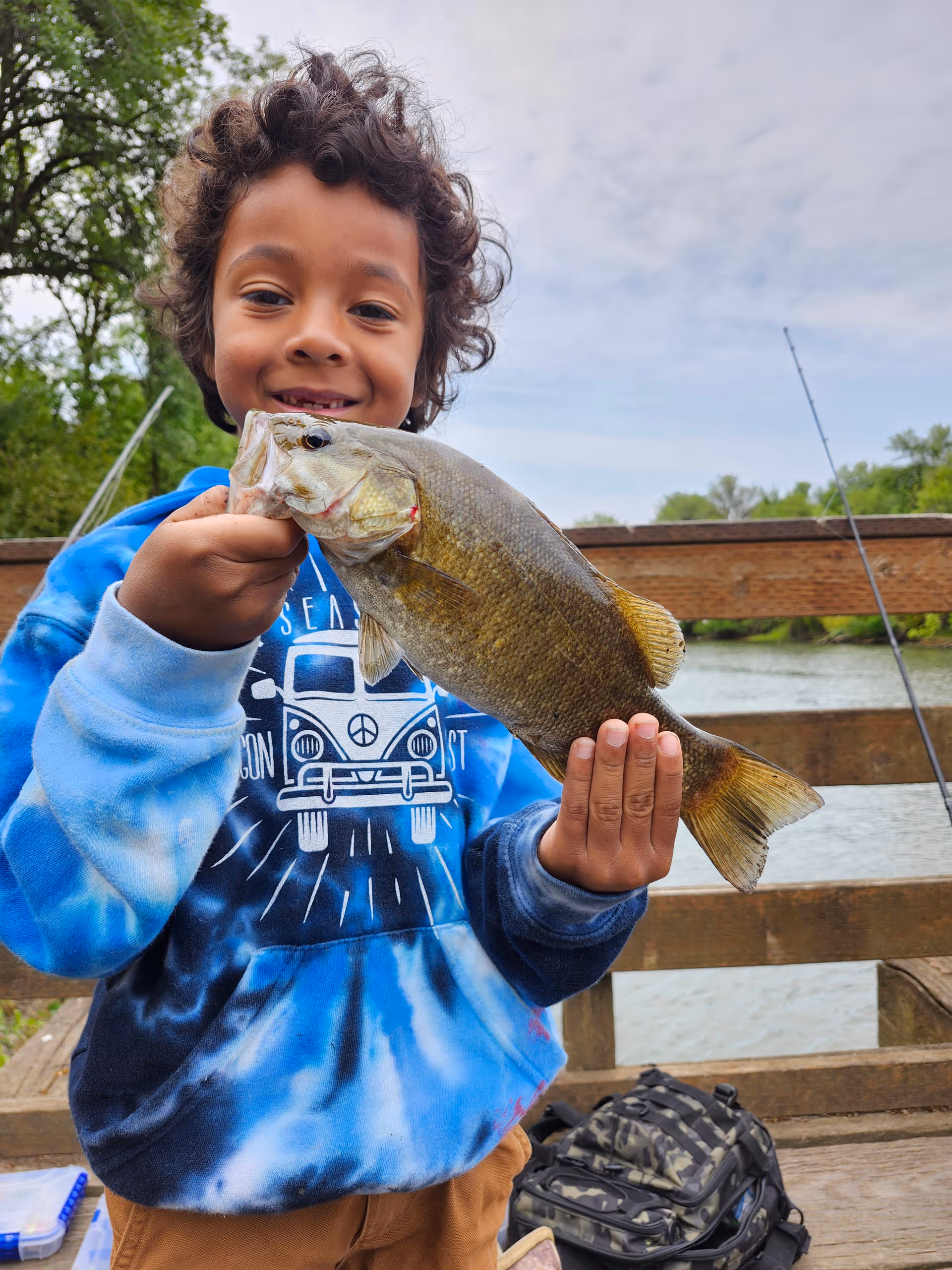 Smiling child holds up a large fish on a wooden dock by a lake.