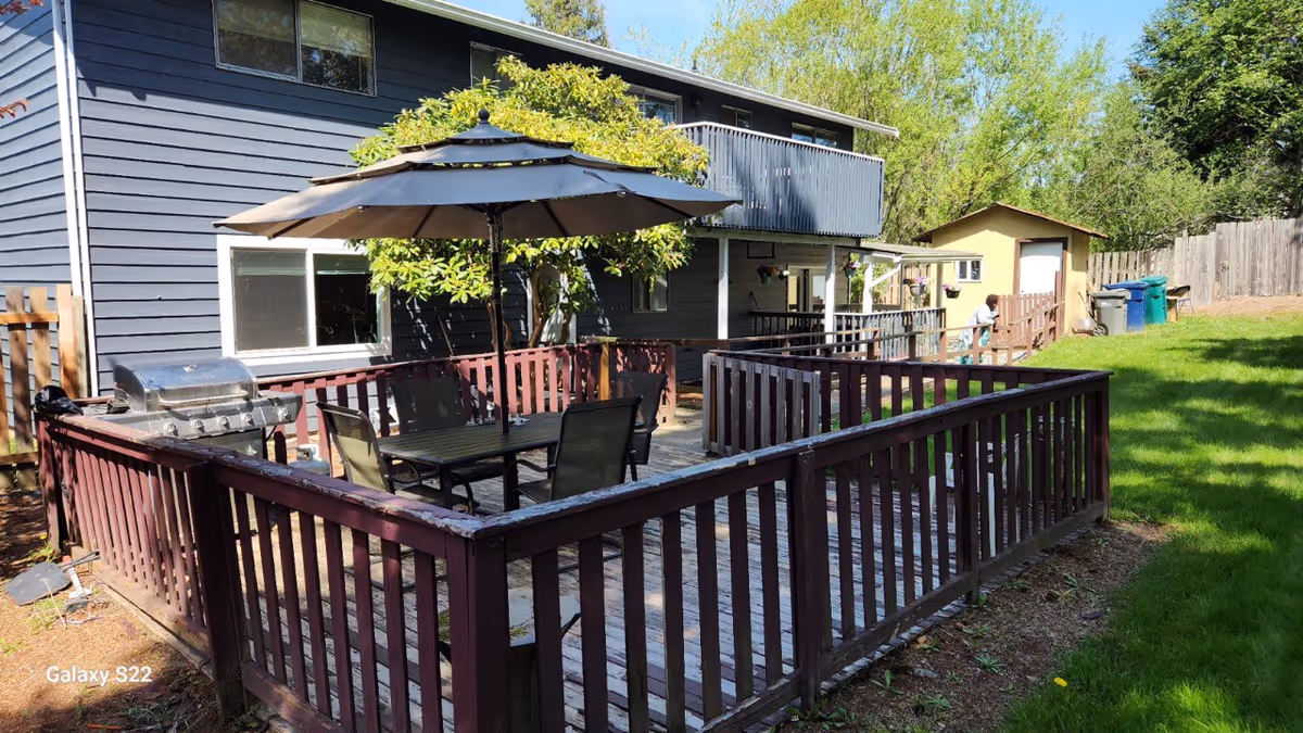 Outdoor wooden deck with table, chairs, umbrella, grill beside a blue house and green lawn.