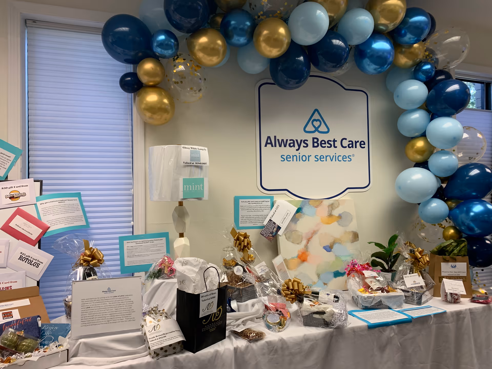 Inside a facility, a blue-gold balloon arch frames an Always Best Care sign above gift tables.