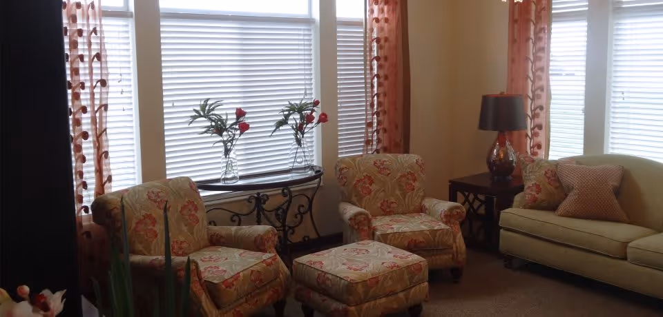 Sunlit sitting area with patterned armchairs, an ottoman, a sofa and side table in front of large windows with blinds and curtains.