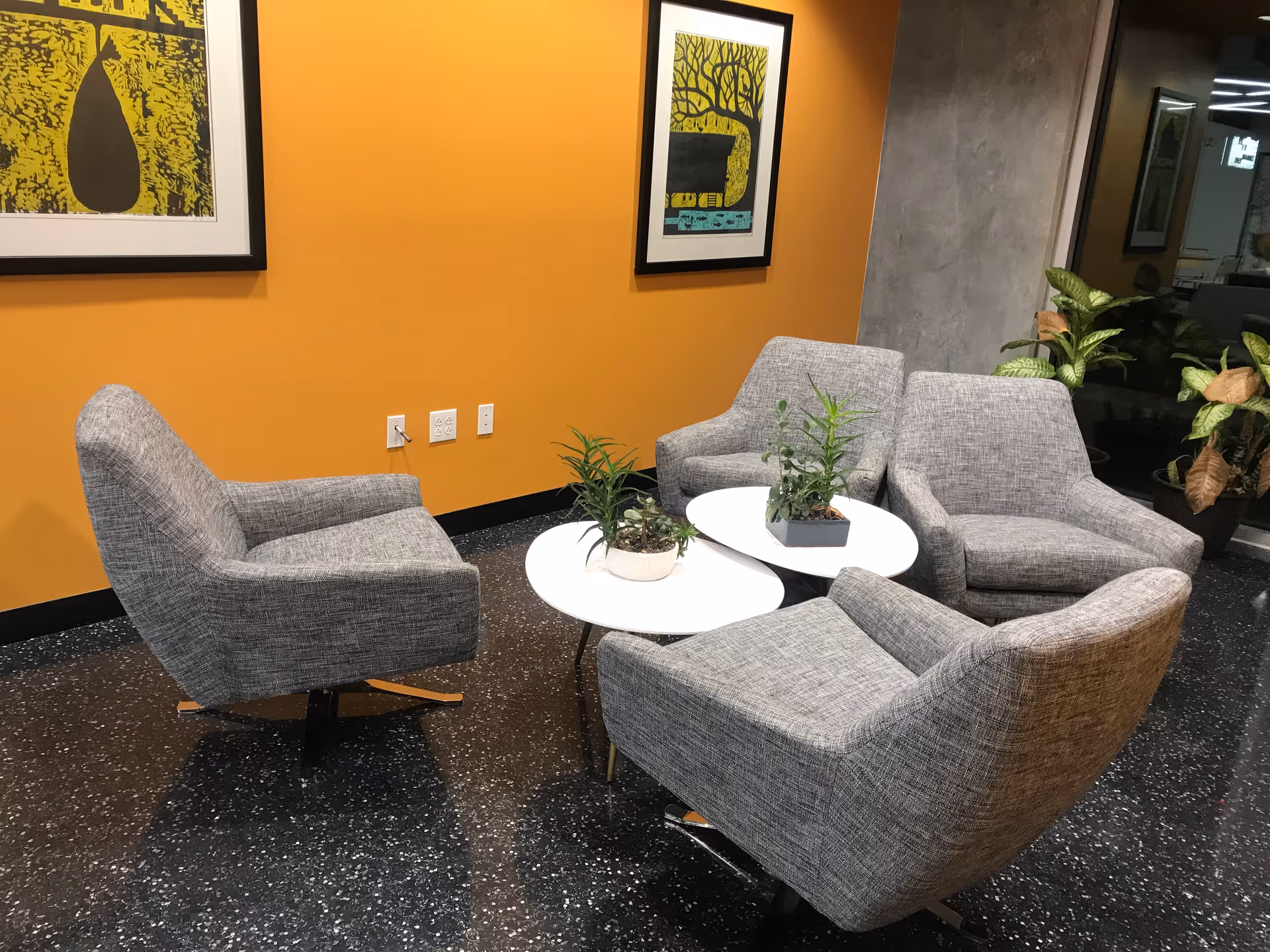 A modern lounge area with four gray armchairs around white round tables near an orange wall.