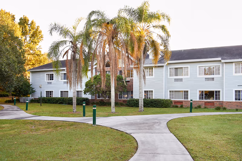 Blue two-story senior living building with a curved concrete walkway and palm trees.