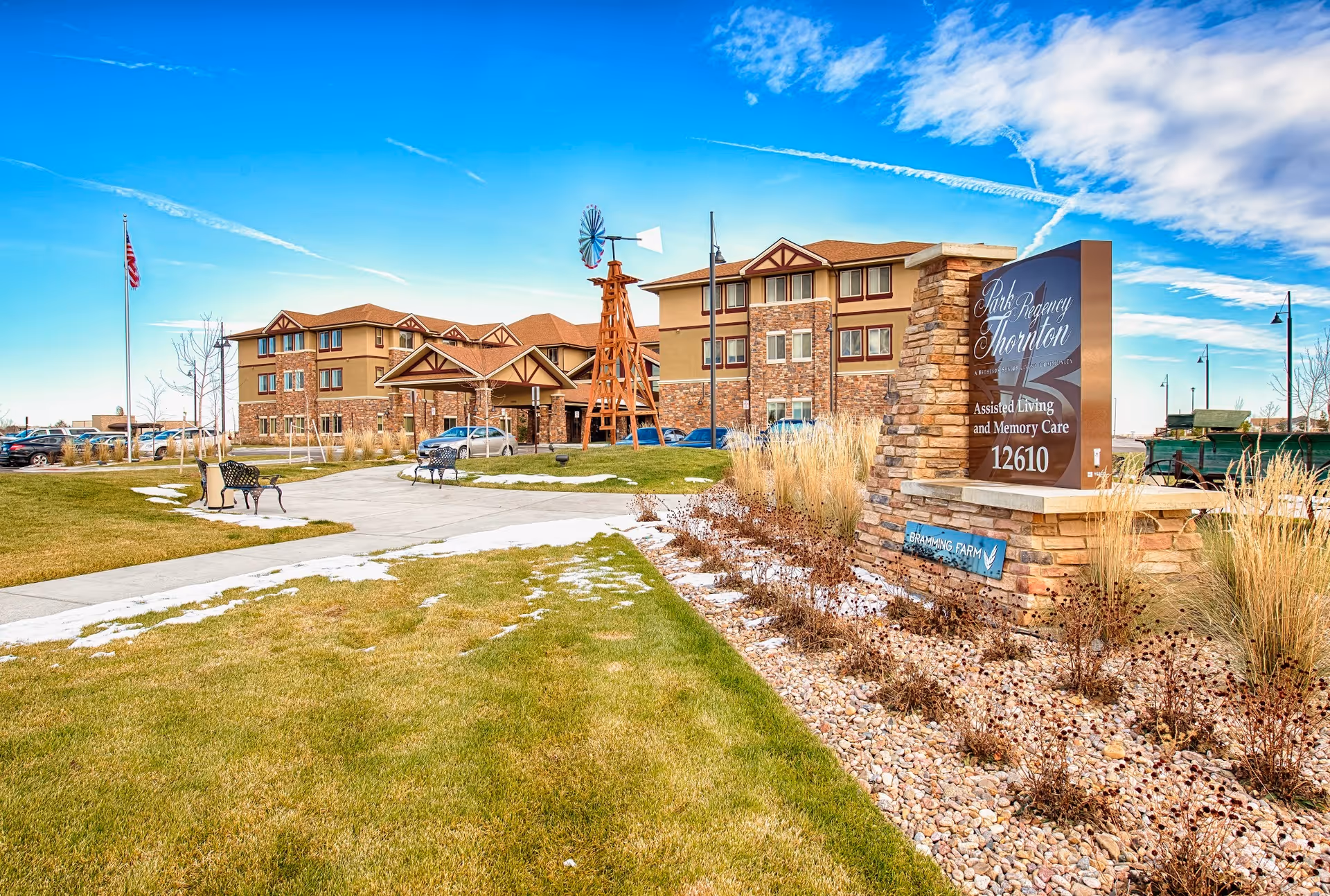 Exterior view of a senior living facility named Park Regency Thornton with a large sign in front, a pathway leading to the entrance, benches, a windmill structure, and a clear blue sky with some clouds.