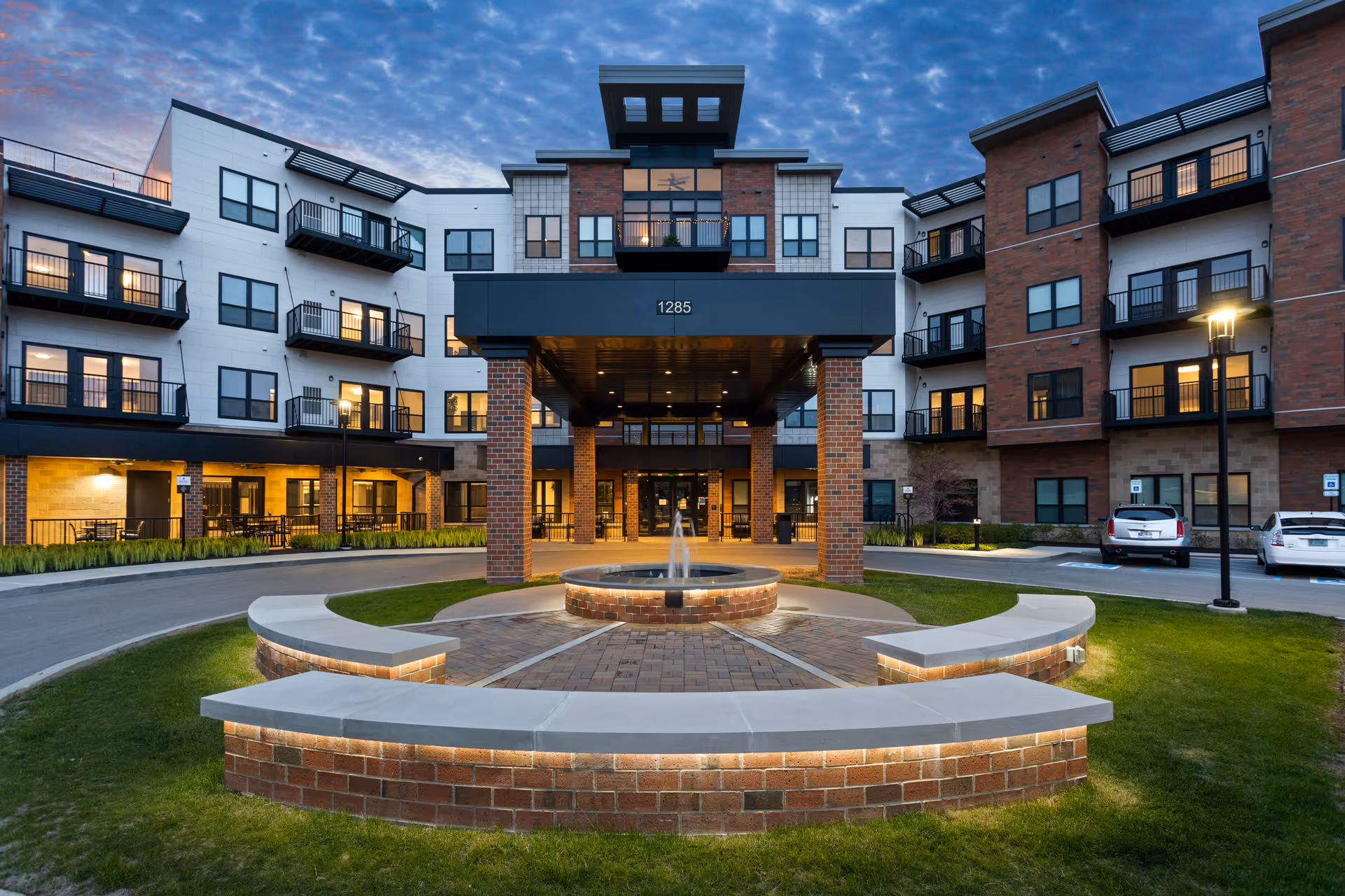 Front entrance of a multi-story senior living building with a circular fountain and curved brick benches lit at dusk.