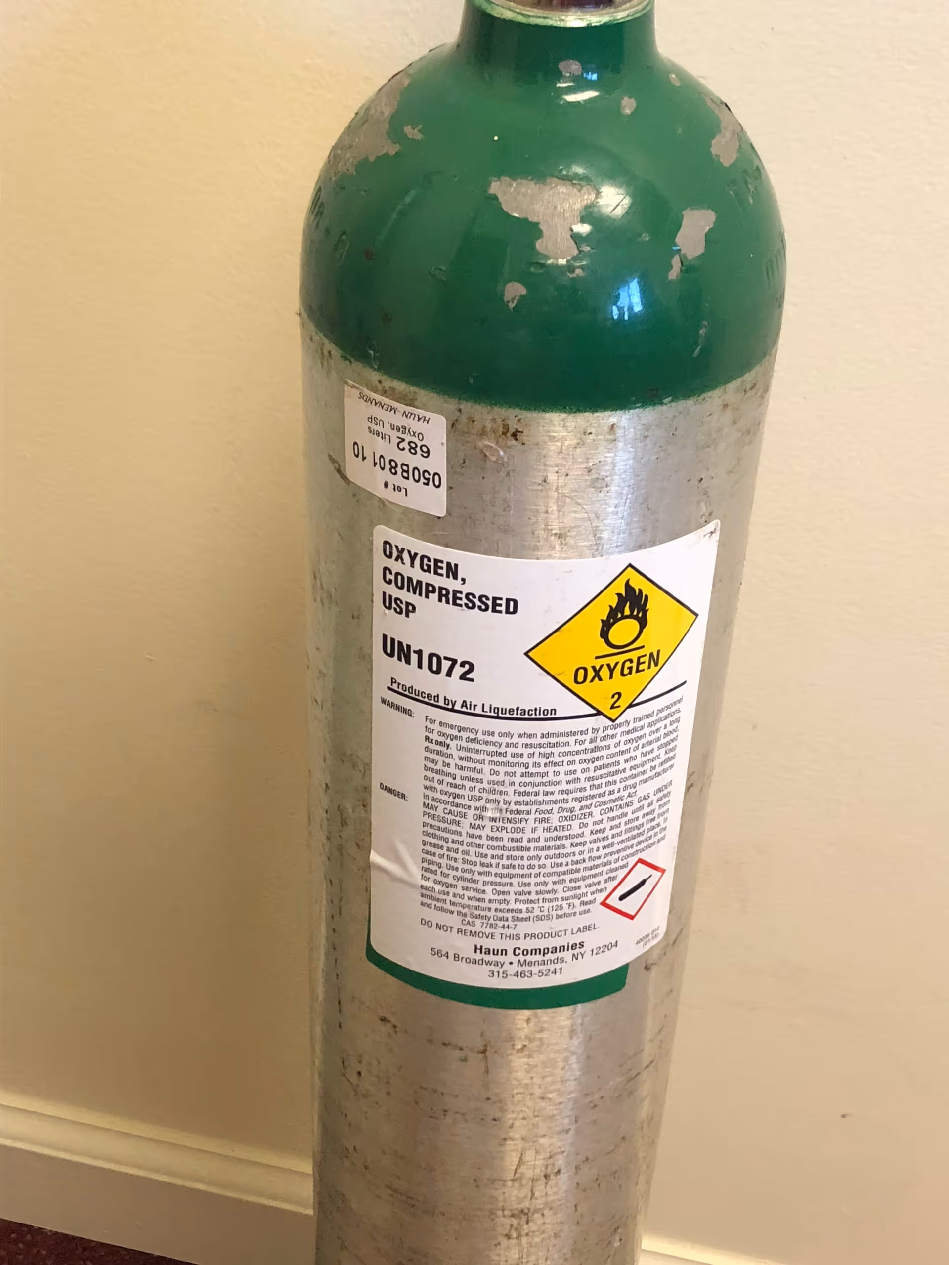 Green oxygen cylinder with a warning label stands indoors against a pale yellow wall.