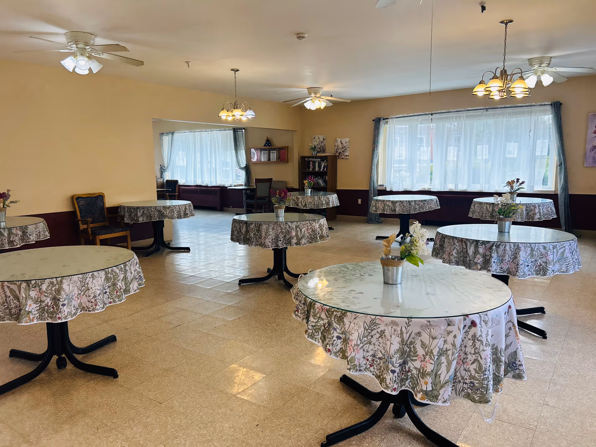 A spacious dining room with several round tables covered with floral tablecloths and small flower arrangements in metal pots. The room has large windows with sheer white curtains, ceiling fans with lights, and beige walls with a dark brown wainscoting. There are a few chairs and a bookshelf in the background.