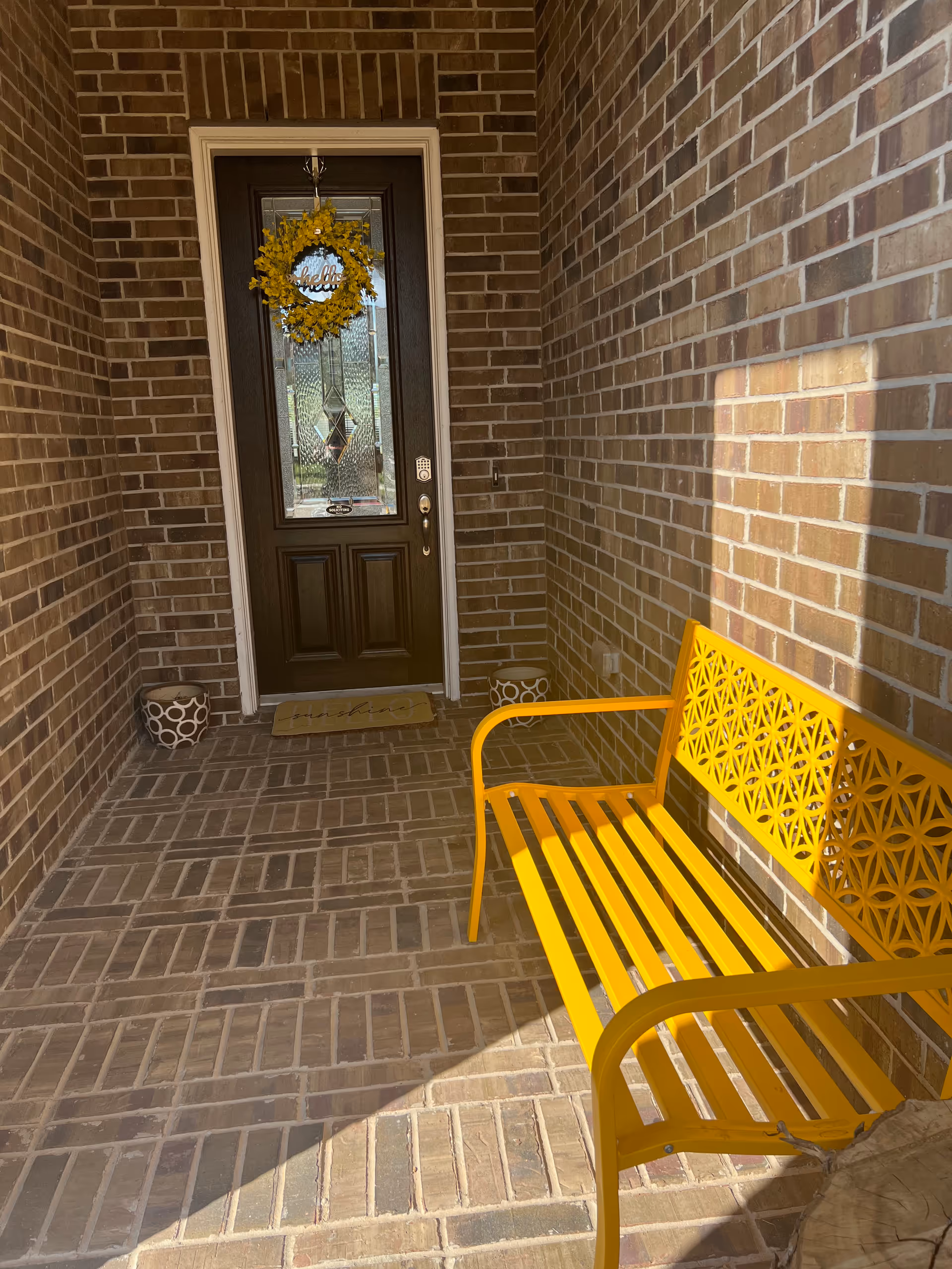 Brick entry porch with a wreath, yellow bench, and potted planters.