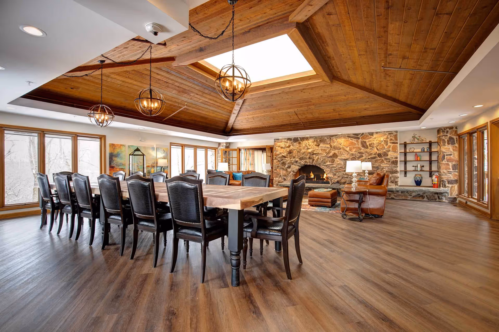 Spacious common room with a long wooden dining table surrounded by chairs, a stone fireplace, leather seating, and a wood-paneled ceiling with a skylight.