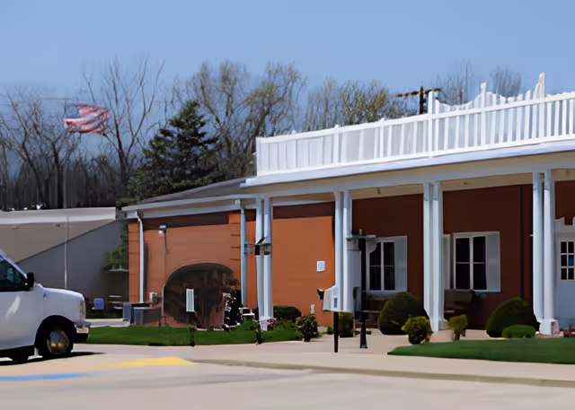 Front exterior of a single-story brick retirement facility with white columns, a covered porch, a parked white van, and an American flag flying nearby.