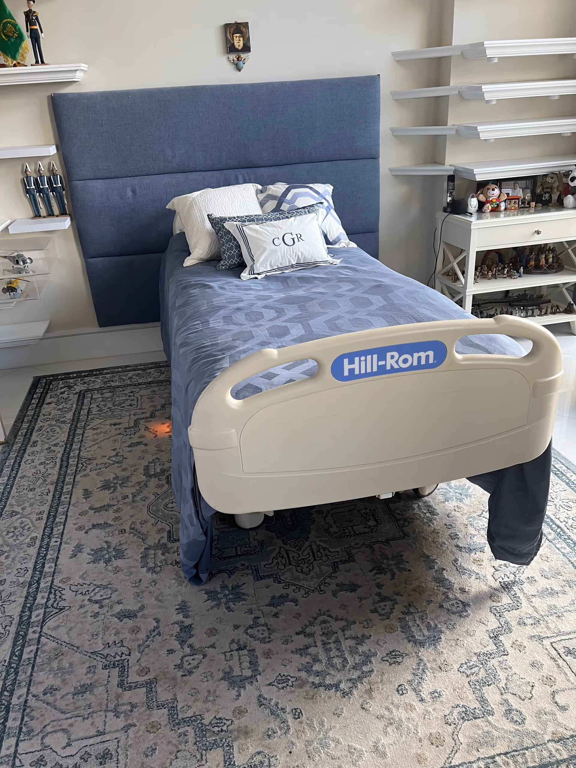 Blue Hill-Rom hospital bed in a resident's bedroom with shelves and decor.