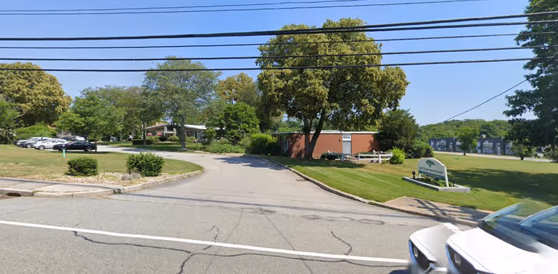 Driveway entrance to a low brick building surrounded by trees and lawns, with a sign on the right and parked cars to the left.