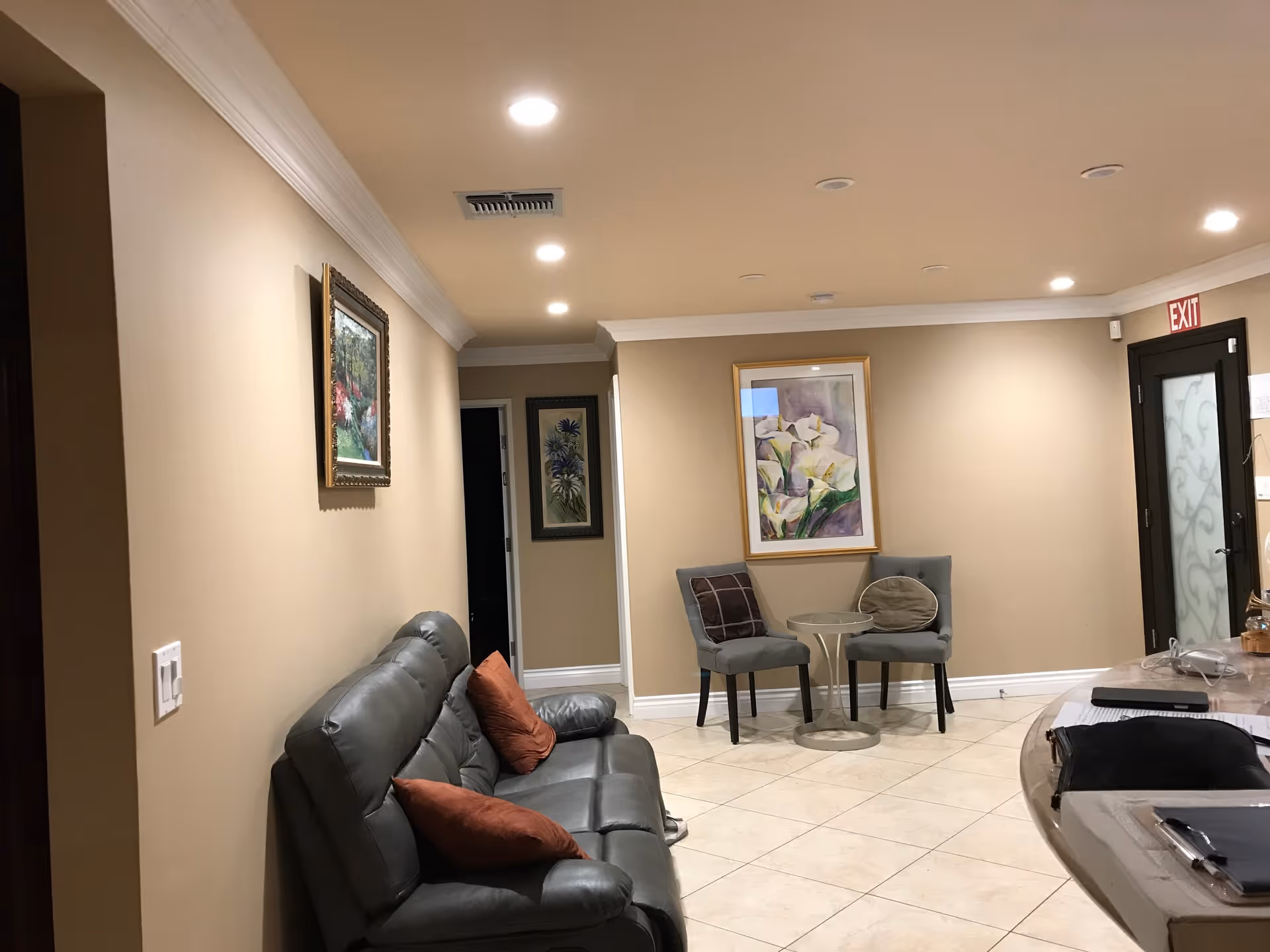 Interior view of a living room area with beige walls and tiled floor. There is a gray leather sofa with two rust-colored pillows on the left side, two gray chairs with cushions around a small round glass table in the center, and framed floral paintings on the walls. Recessed ceiling lights illuminate the space, and a door with frosted glass and an exit sign is visible on the right side.