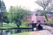 Pink brick senior living facility across a pond with a wooden footbridge and trees.