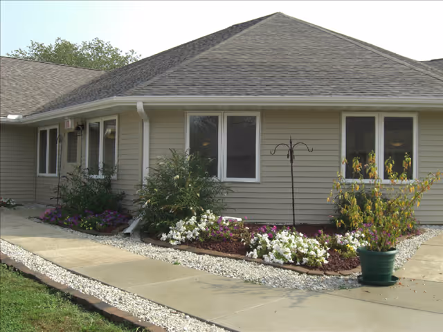 Single-story beige-sided building with a sloped roof, a paved walkway, and landscaped flowerbeds in front.