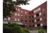 Red brick senior living building with multiple windows, greenery, and a green striped awning entrance.
