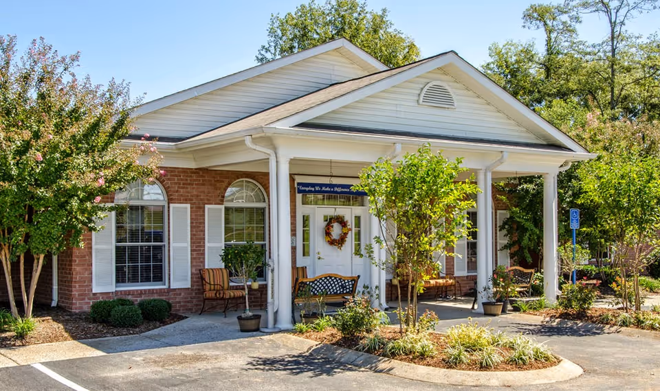 Front exterior view of a single-story brick building with white trim and a covered entrance supported by white columns. There are benches and potted plants near the entrance, surrounded by landscaped bushes and trees under a clear blue sky.