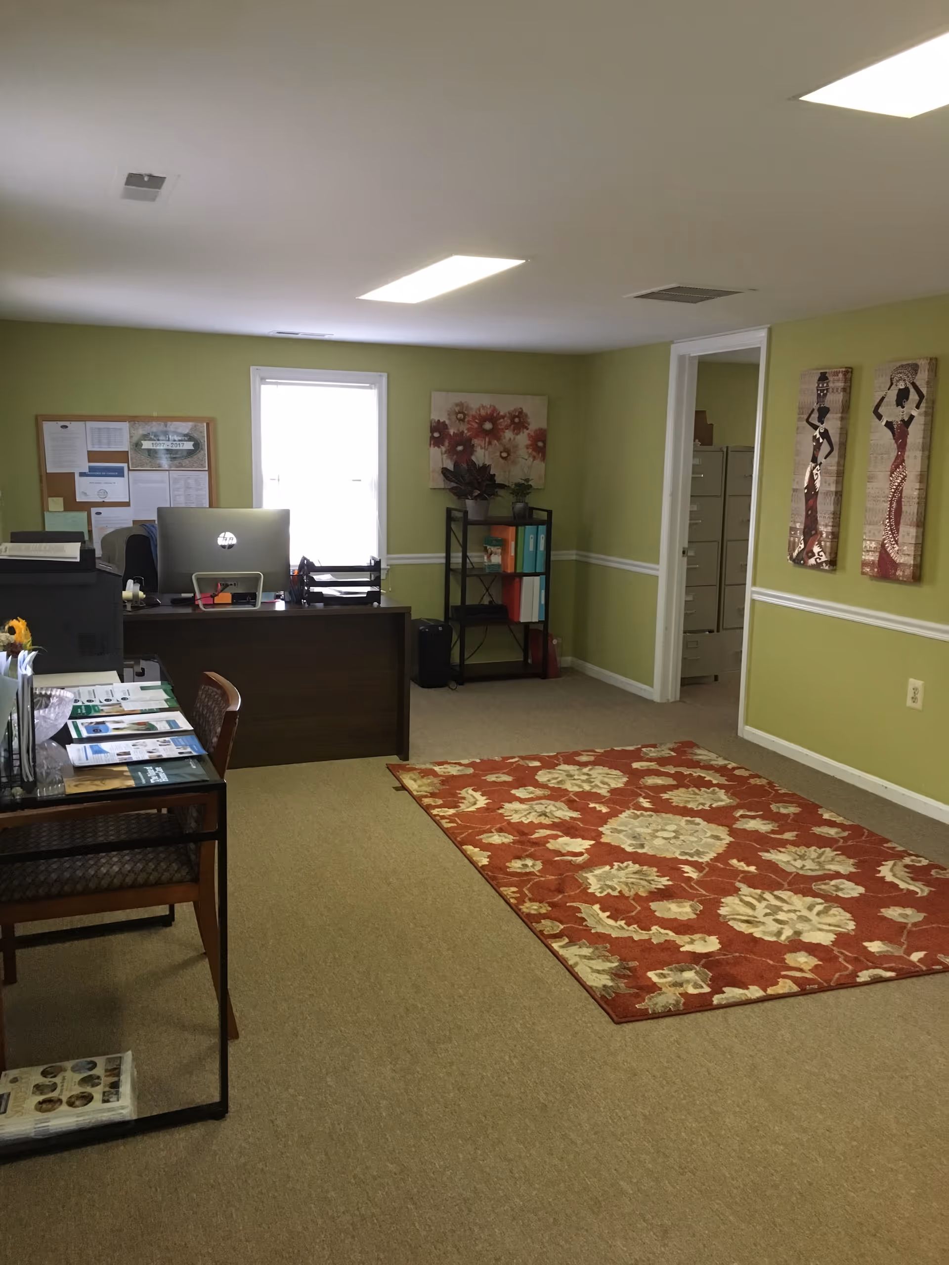 Office reception area with desk, computer, bulletin board, bookshelf, and green walls.