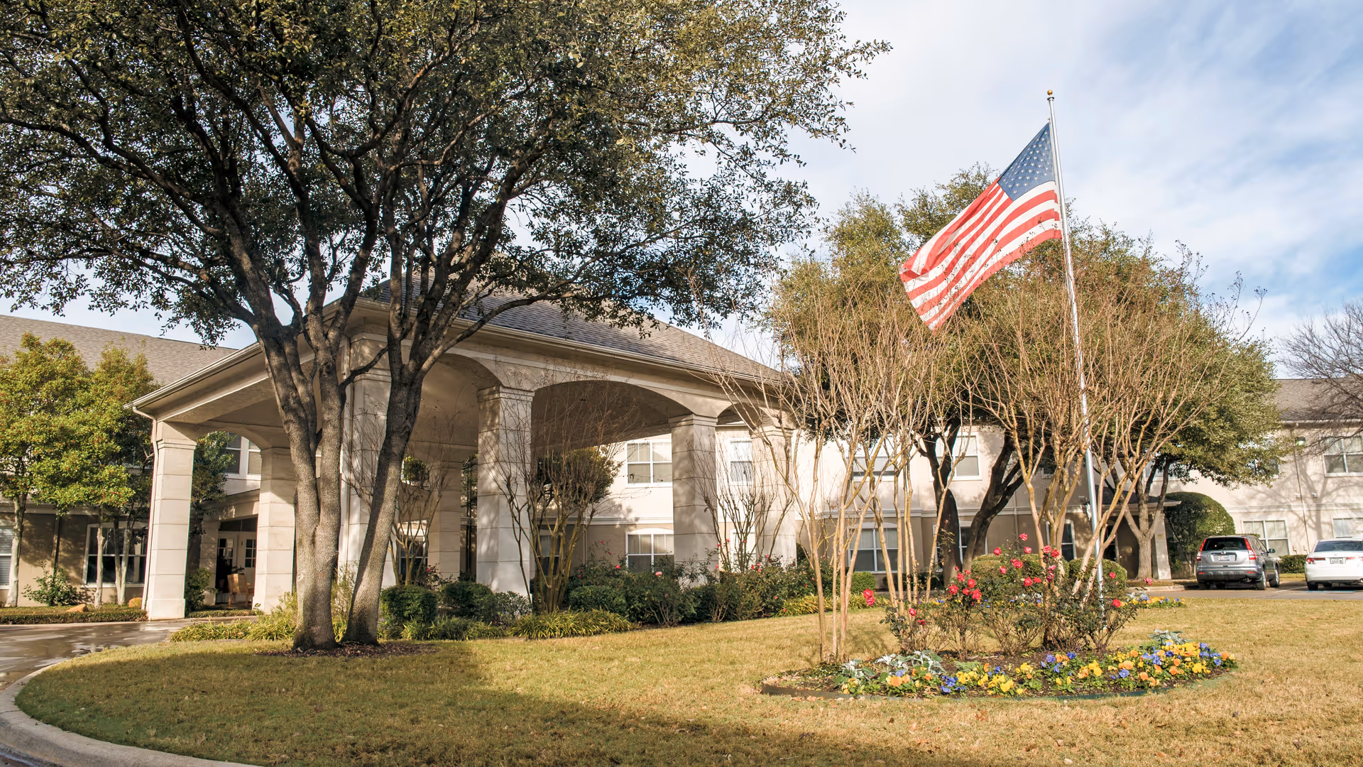 Brookdale Eden Estates exterior: entrance with portico, American flag, trees, and landscaped flower bed.
