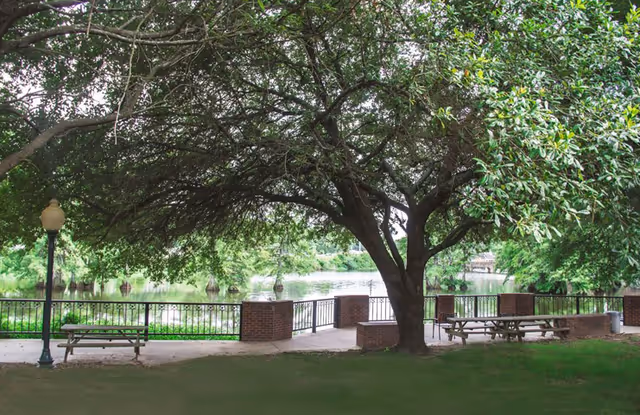 A peaceful outdoor area with large trees providing shade over picnic tables and benches near a body of water, surrounded by a black metal fence with brick pillars.