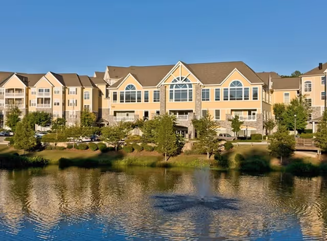 Multi-story beige senior living building with large arched windows and balconies reflected across a pond with a small fountain.