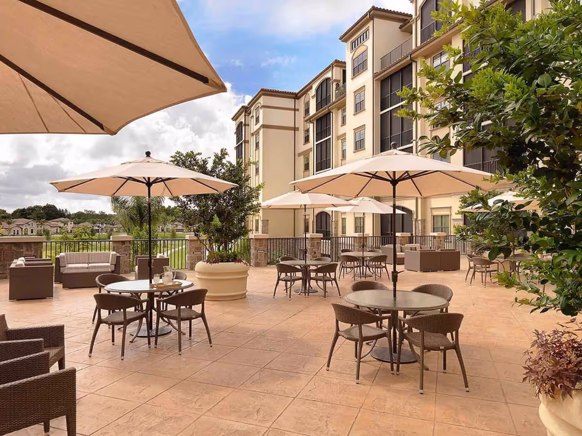 Outdoor terrace with tables, chairs, umbrellas, and greenery beside a multi-story senior living building.