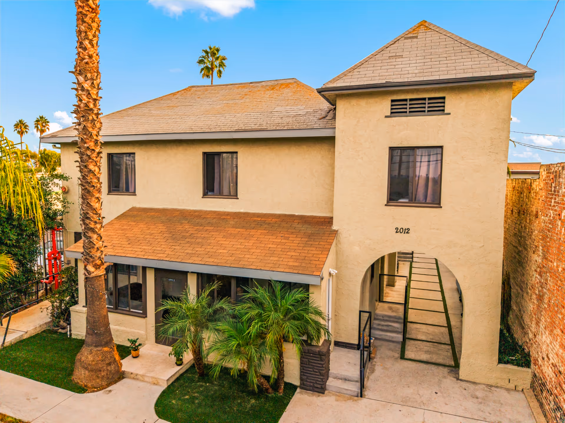 Two-story beige building with palm trees, arched entrance, and courtyard.