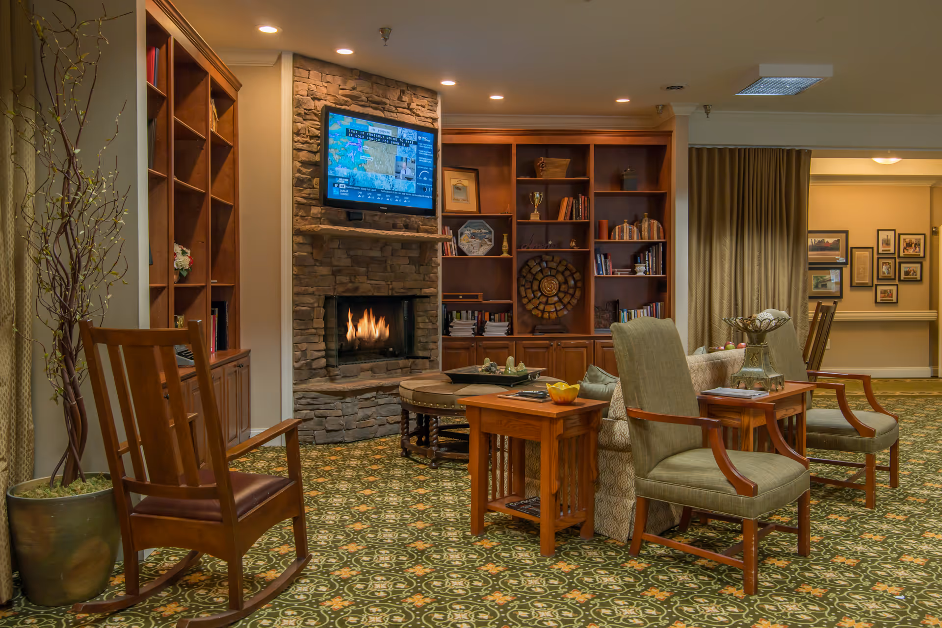 A cozy living room area in a senior living facility featuring a stone fireplace with a flat-screen TV mounted above it, wooden bookshelves filled with books and decorative items, a patterned green carpet, a wooden rocking chair, two upholstered armchairs, and wooden side tables with decorative bowls and books. The room is warmly lit with recessed ceiling lights.