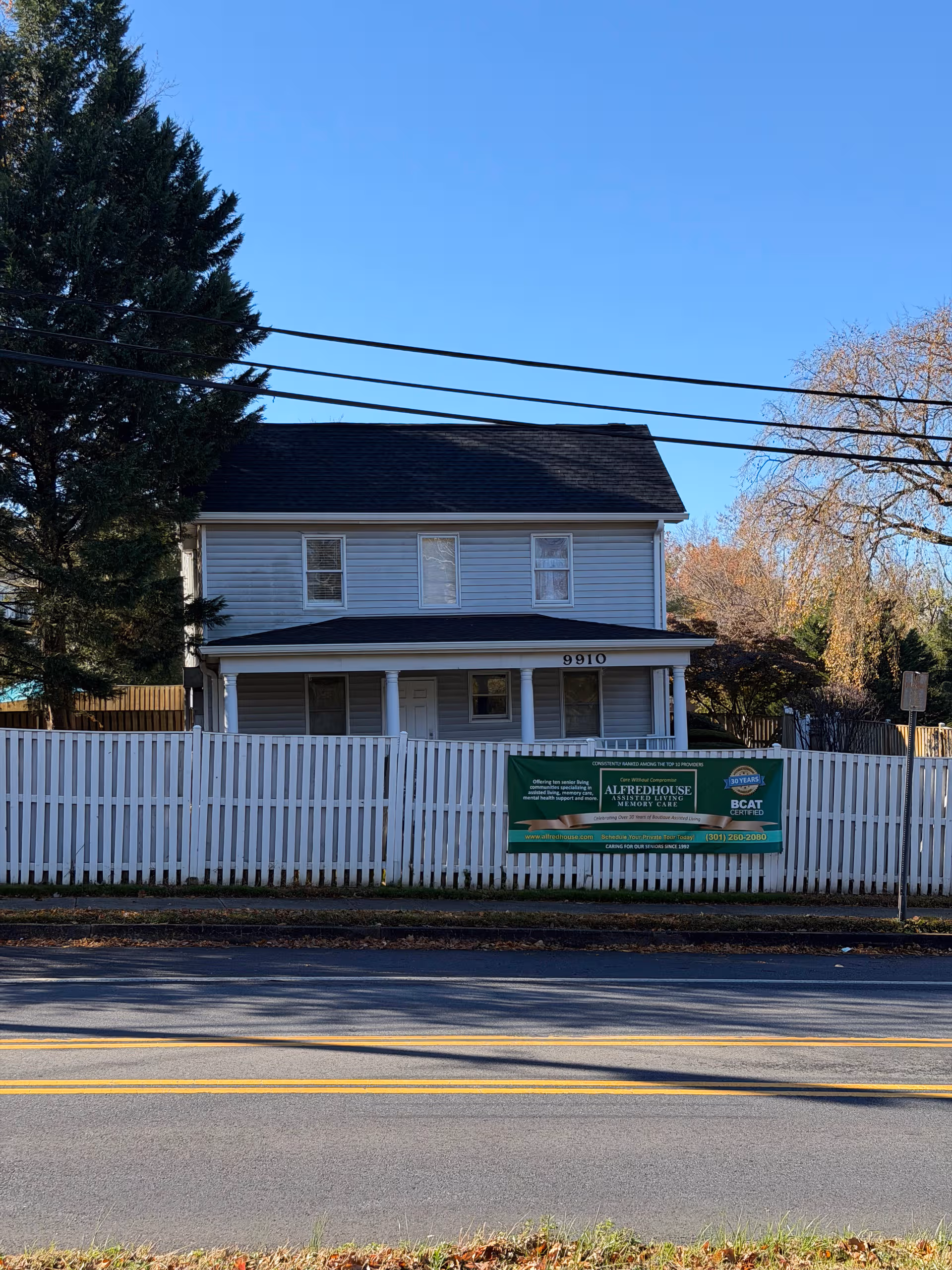Two-story gray house with a white picket fence and banner in front.
