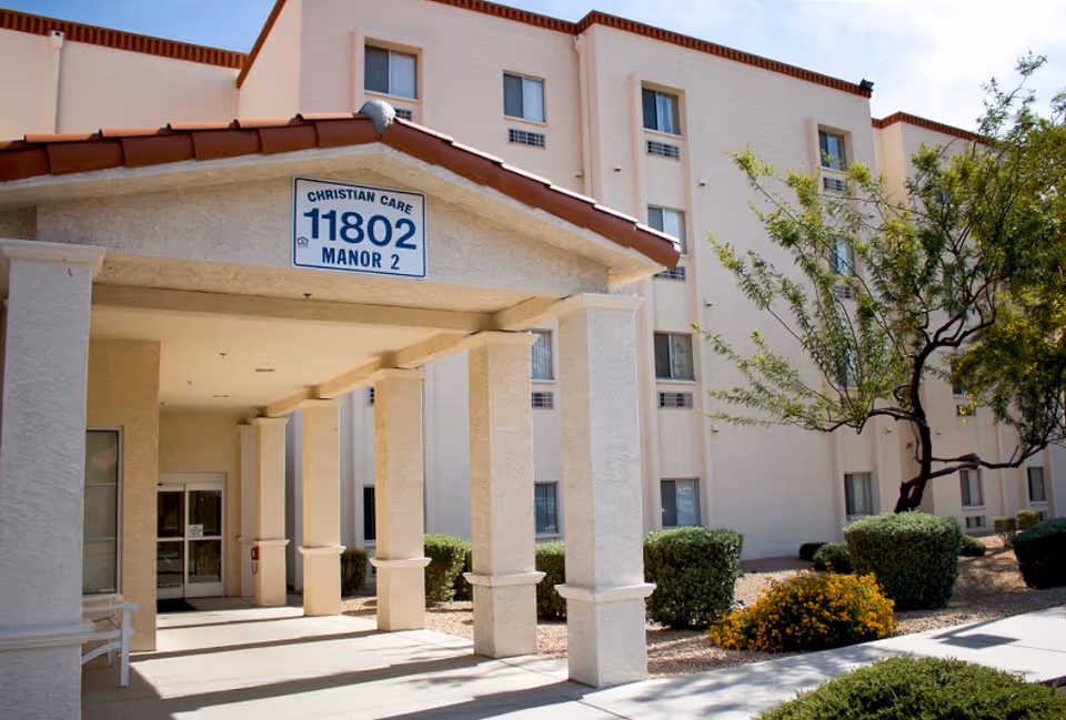 Front entrance canopy and facade of an assisted-living building with a sign reading "Christian Care 11802 Manor 2" and surrounding shrubs.