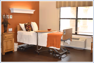 A senior living bedroom with an adjustable hospital-style bed, nightstand, window and warm orange accent wall.
