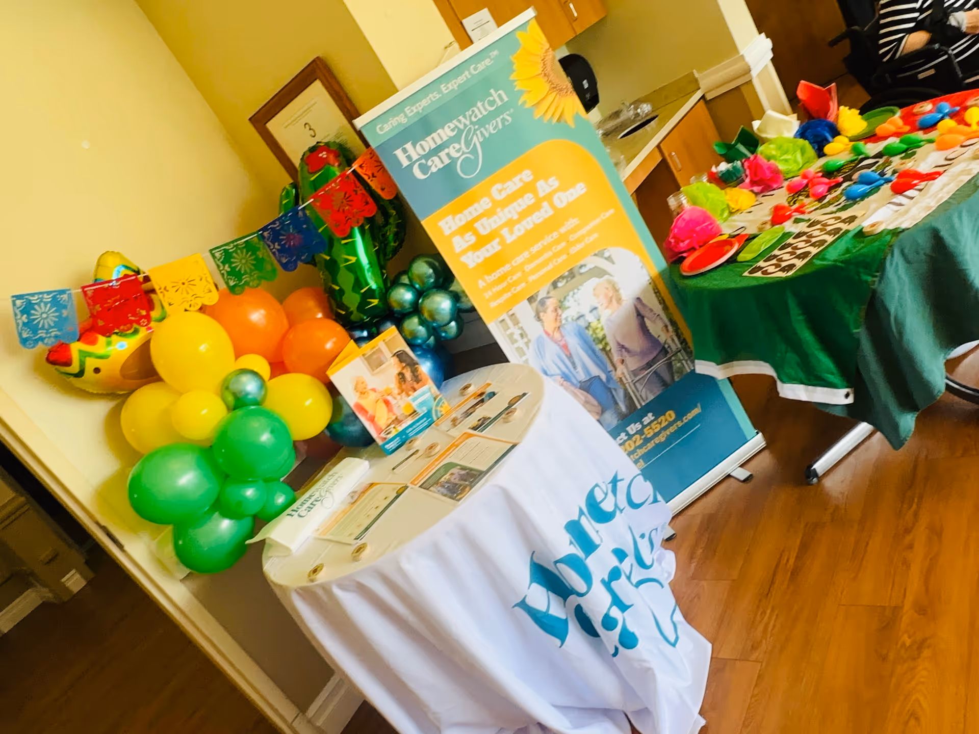 Indoor event area with balloons, tables, and a Homewatch CareGivers banner.